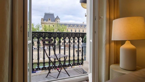 A room with a view of a historic building across a canal, a balcony with a chair, and a lamp on a bedside table. - Le Balcon de Notre Dame (Paris)