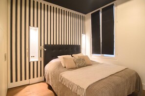 A bedroom with a striped wall, a black headboard, and a bed with a beige blanket and pillows. - Le Balcon de Notre Dame (Paris)
