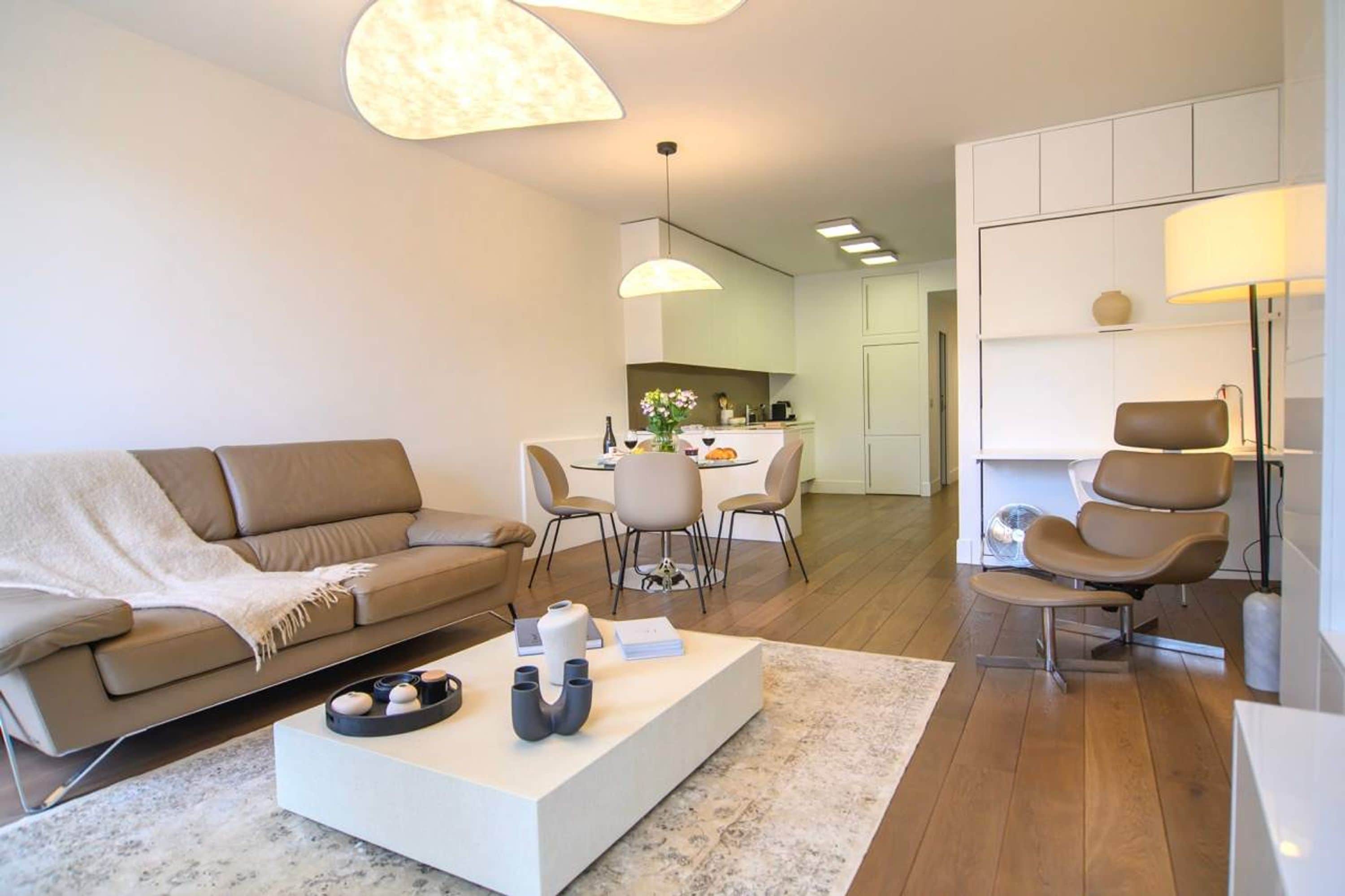 A modern living room with a sofa, coffee table, and chairs. There is a dining area with a table and chairs in the background.