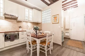Eine Küche mit weißen Schränken, einem Holzespeisetisch und Stühlen. Es gibt ein Fenster mit Jalousien, ein Bild an der Wand und eine Pendelleuchte.