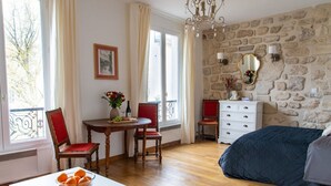 A bedroom with a stone wall, a bed, a dresser, a desk with a chair, and a window with curtains. - Studio en Isle - Monthly Rental (Paris)