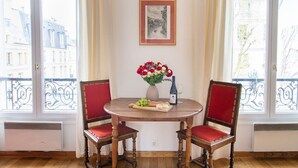 A dining area with a round wooden table, two red upholstered chairs, a vase with flowers, a bottle of wine, and a plate with grapes and cheese. - Studio en Isle - Monthly Rental (Paris)