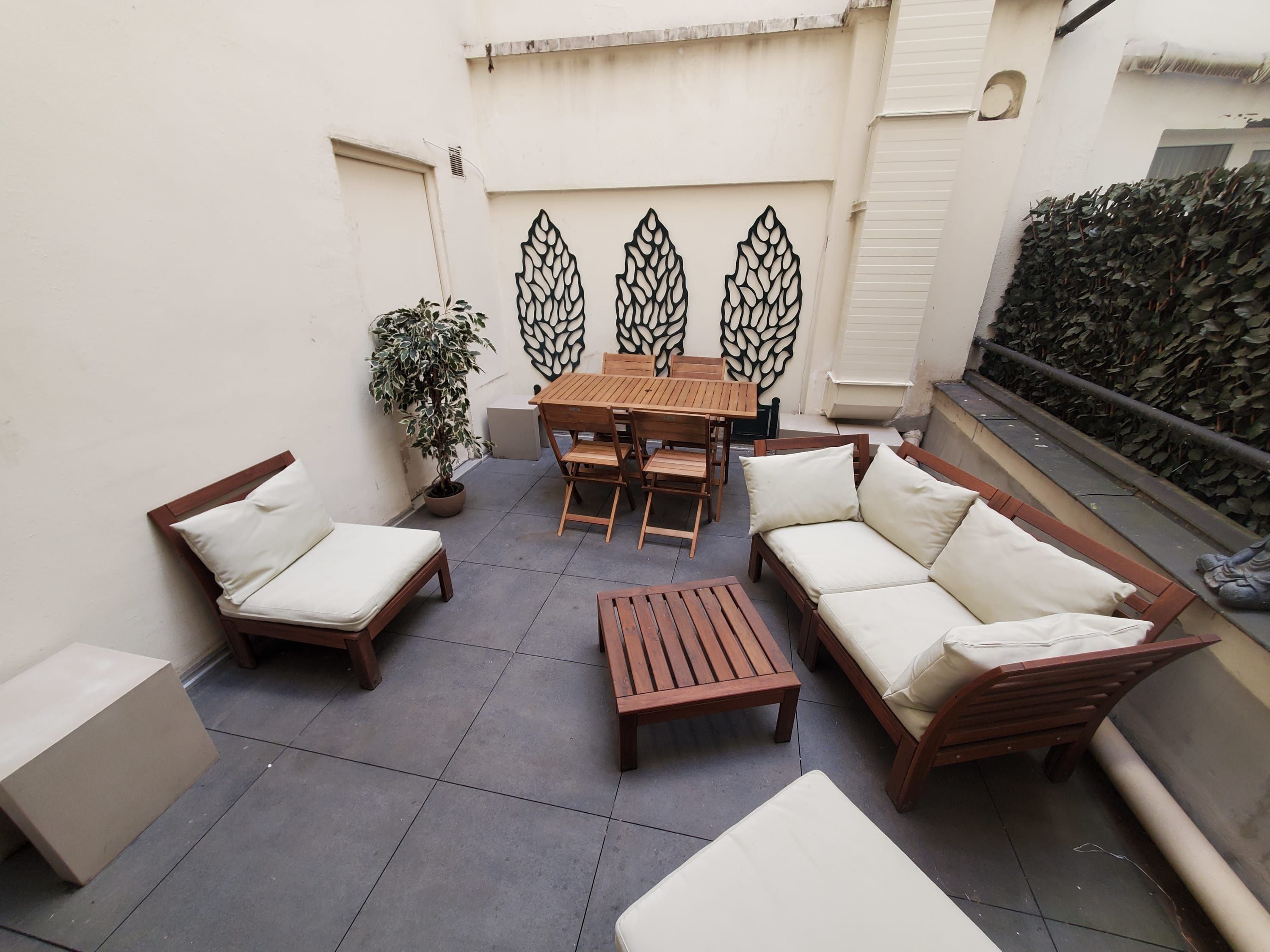 A patio with a wooden table, chairs, and a sofa. There is a potted plant and a decorative wall panel.