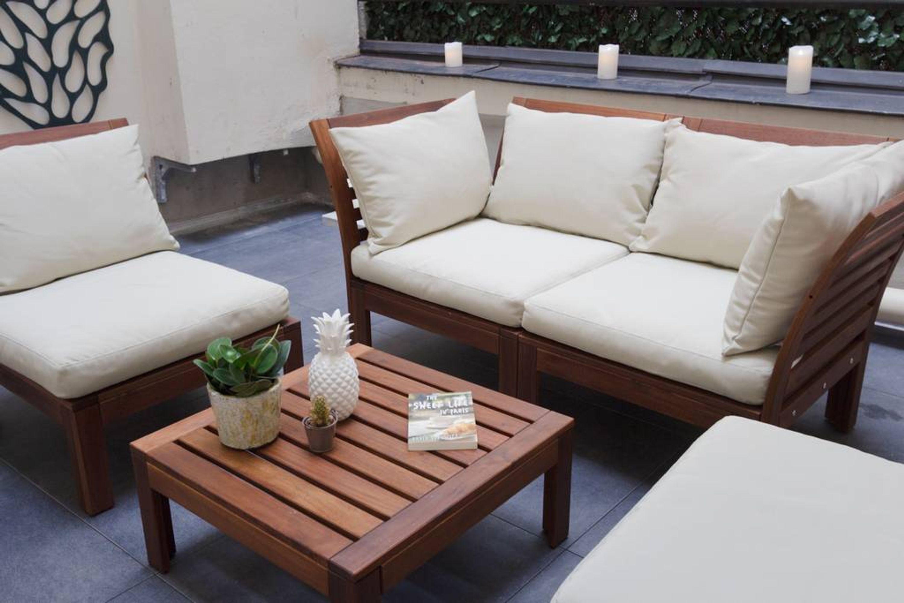 Un espace de détente extérieur en bois, avec un canapé et deux fauteuils, une petite table agrémentée d’une plante et d’un ananas, et des bougies disposées sur une étagère.