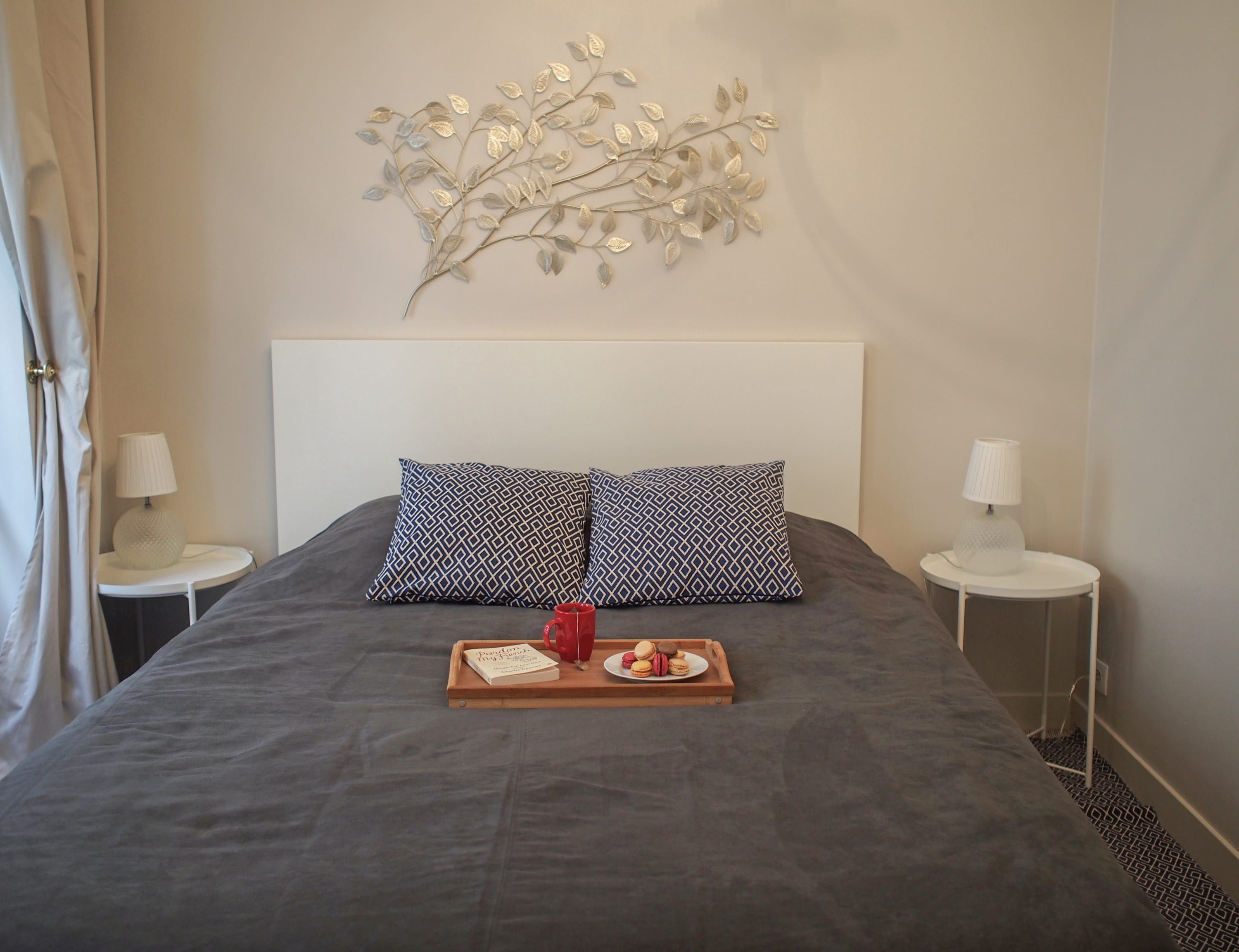 Un lit bien fait, avec un plateau de nourriture, deux coussins à motifs et une œuvre d’art accrochée au mur.