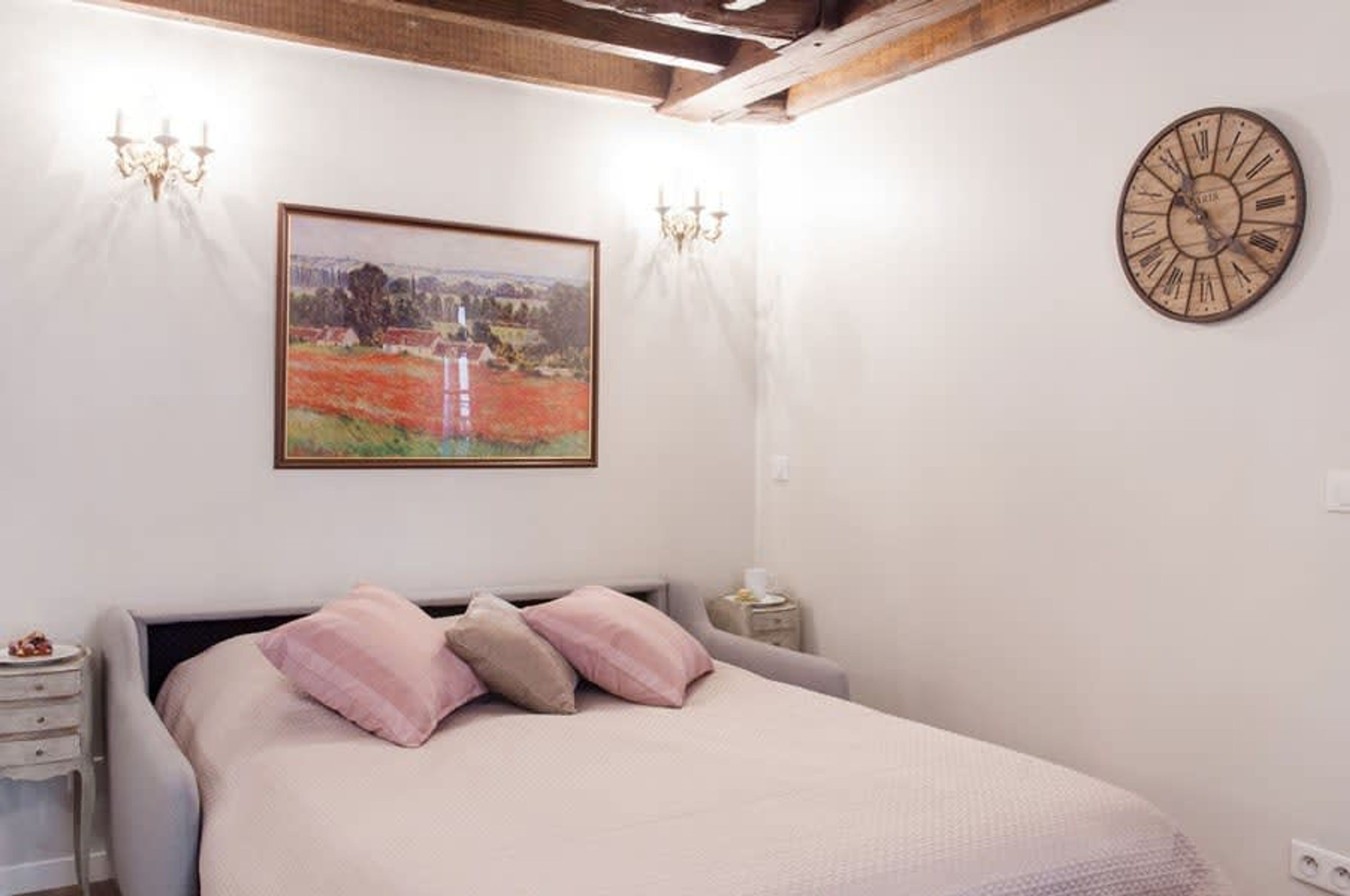 Quarto com cama, um quadro, relógio de parede e duas arandelas.