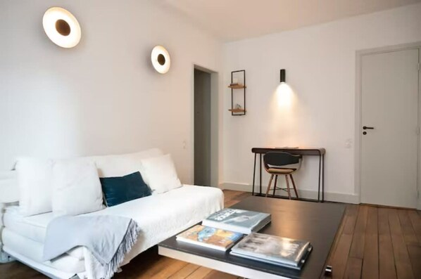 A modern, minimalist room with a white sofa, a small desk with a chair, and a door. - Villa Montparnasse - 15th arr (Paris)
