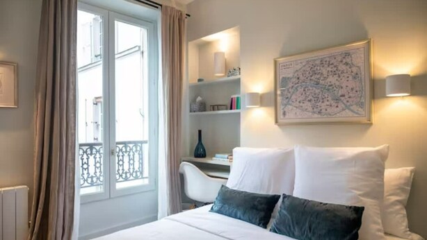 A hotel room with a bed, a desk, a chair, a window with curtains, a map on the wall, and a shelf with books and a vase. - Villa Montparnasse - 15th arr (Paris)