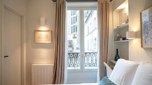 A hotel room with a bed, a radiator, a window with curtains, a view of buildings, and a shelf with items. - Villa Montparnasse - 15th arr (Paris)