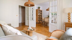 A living room with a sofa, a round coffee table, and a wicker chair. There is a wooden cabinet and a dining table with chairs in the background.