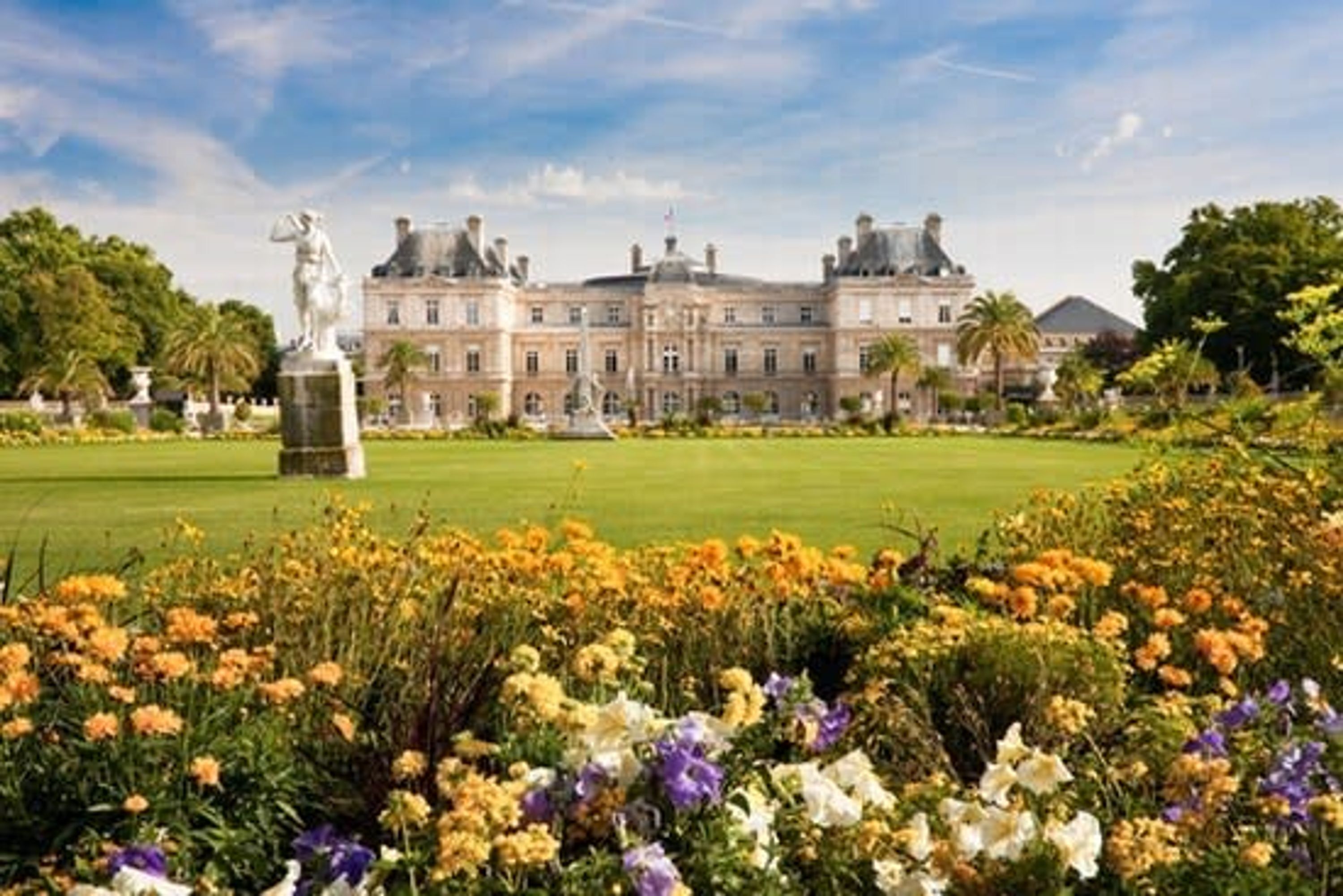 A large, historic building with a statue in front, surrounded by a well-maintained garden with various flowers.