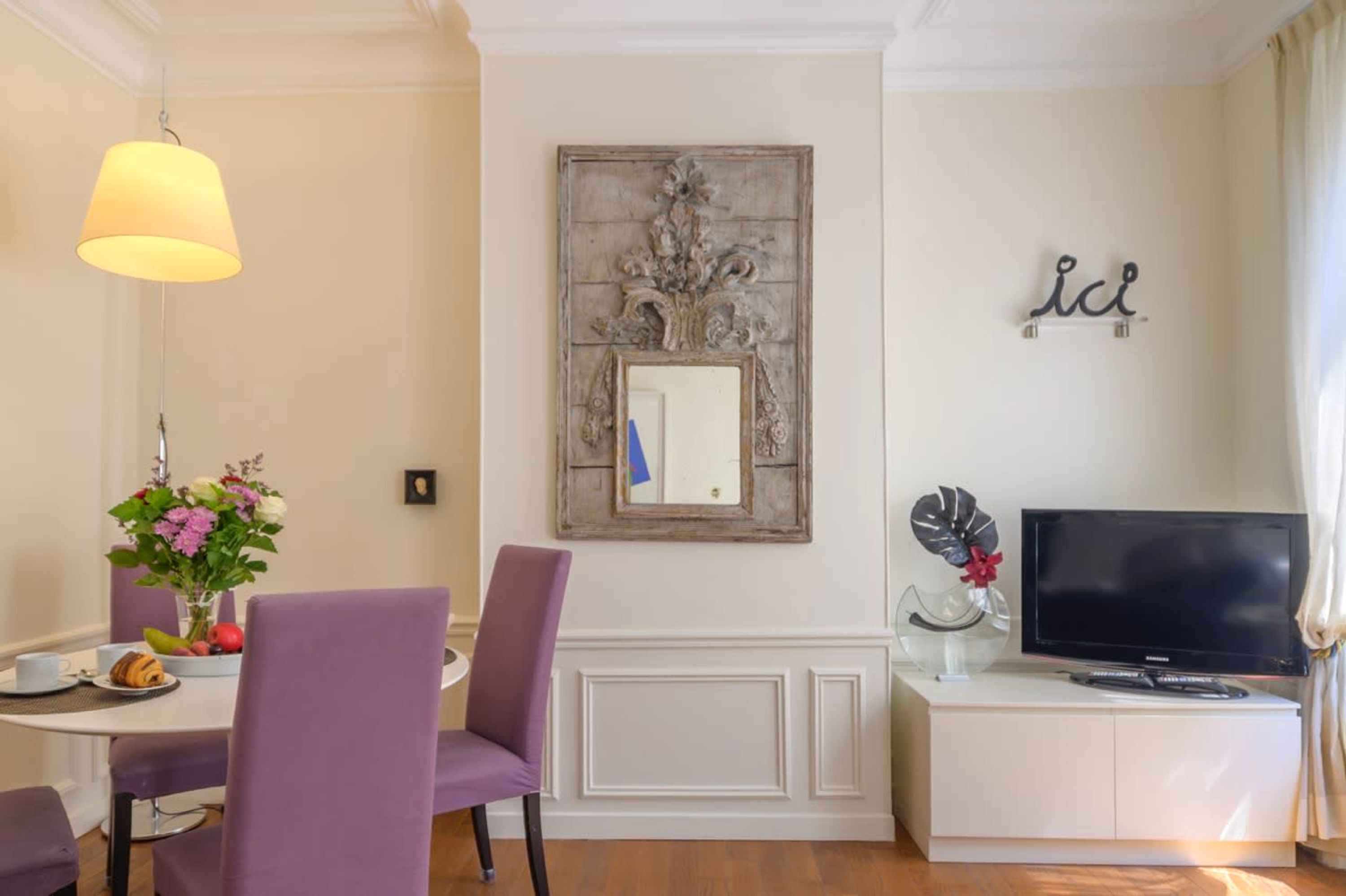 A dining area with a table, chairs, a lamp, a television, and a decorative mirror.