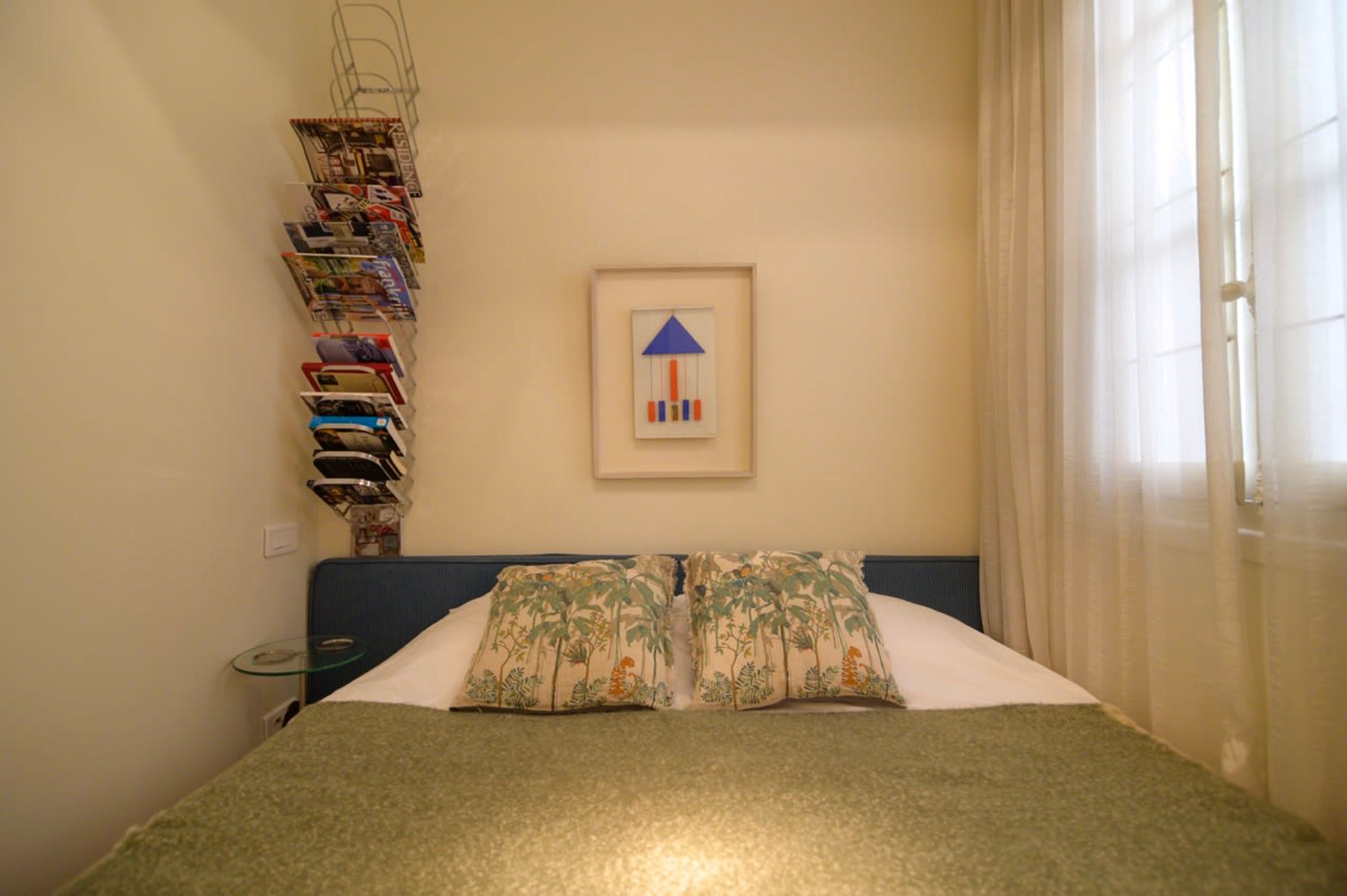 Une chambre d’hôtel bien rangée, avec un lit, des oreillers, un tableau encadré et une étagère fixée au mur où sont posés des magazines.