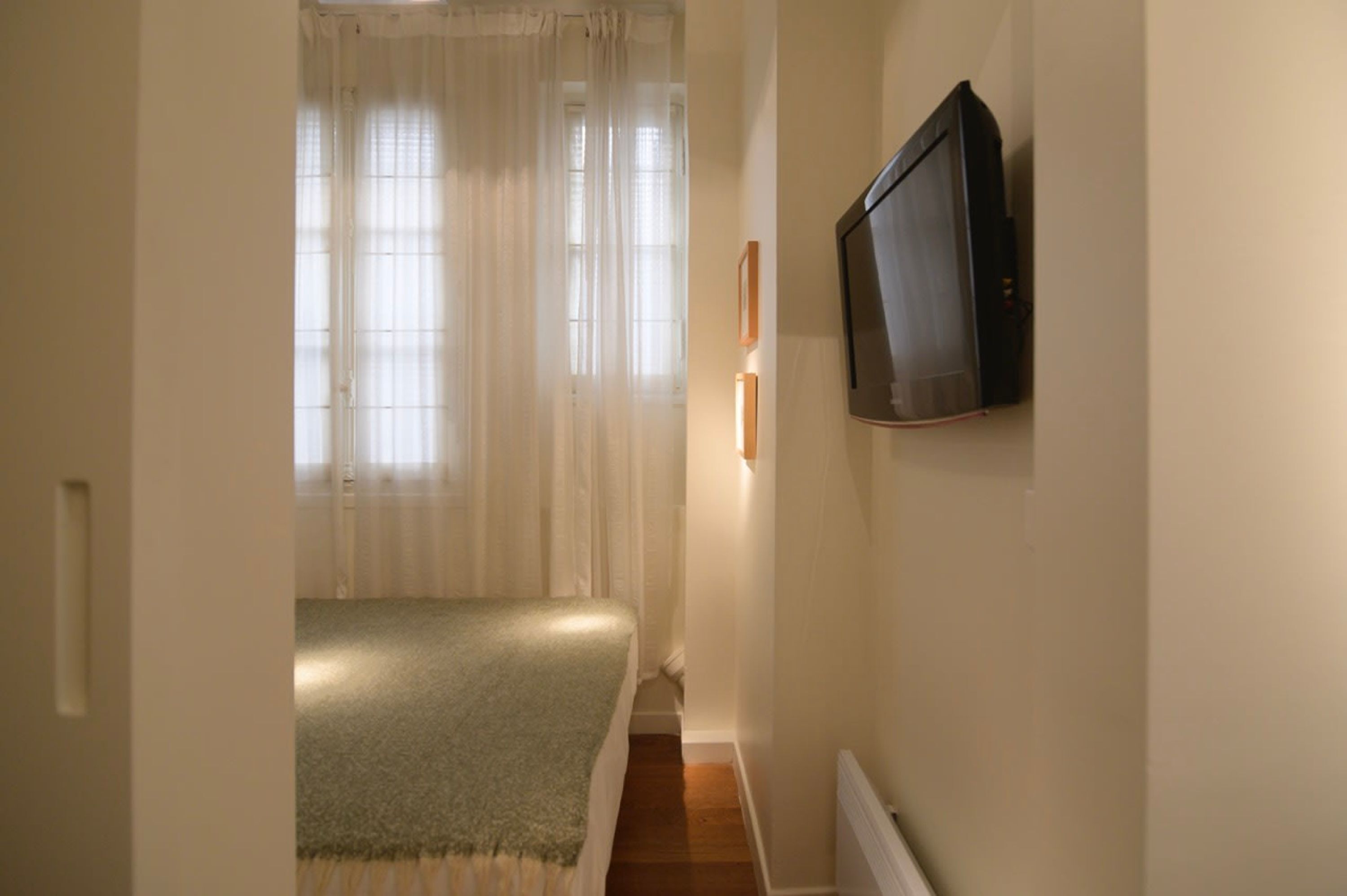 Un couloir étroit, avec un lit au fond, un téléviseur fixé au mur et une fenêtre ornée de rideaux légers.