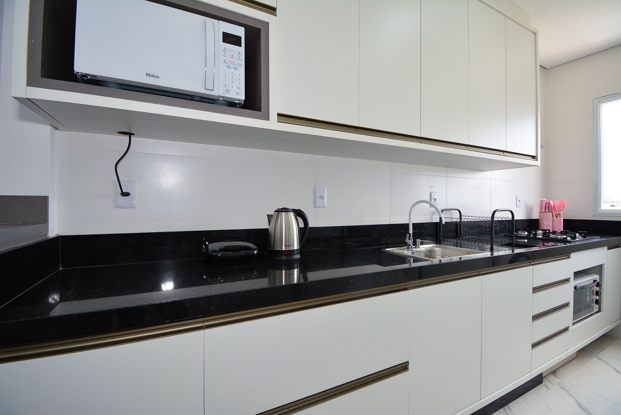 Una cocina moderna con armarios blancos, encimera negra, microondas integrado, hervidor y lavabo.
