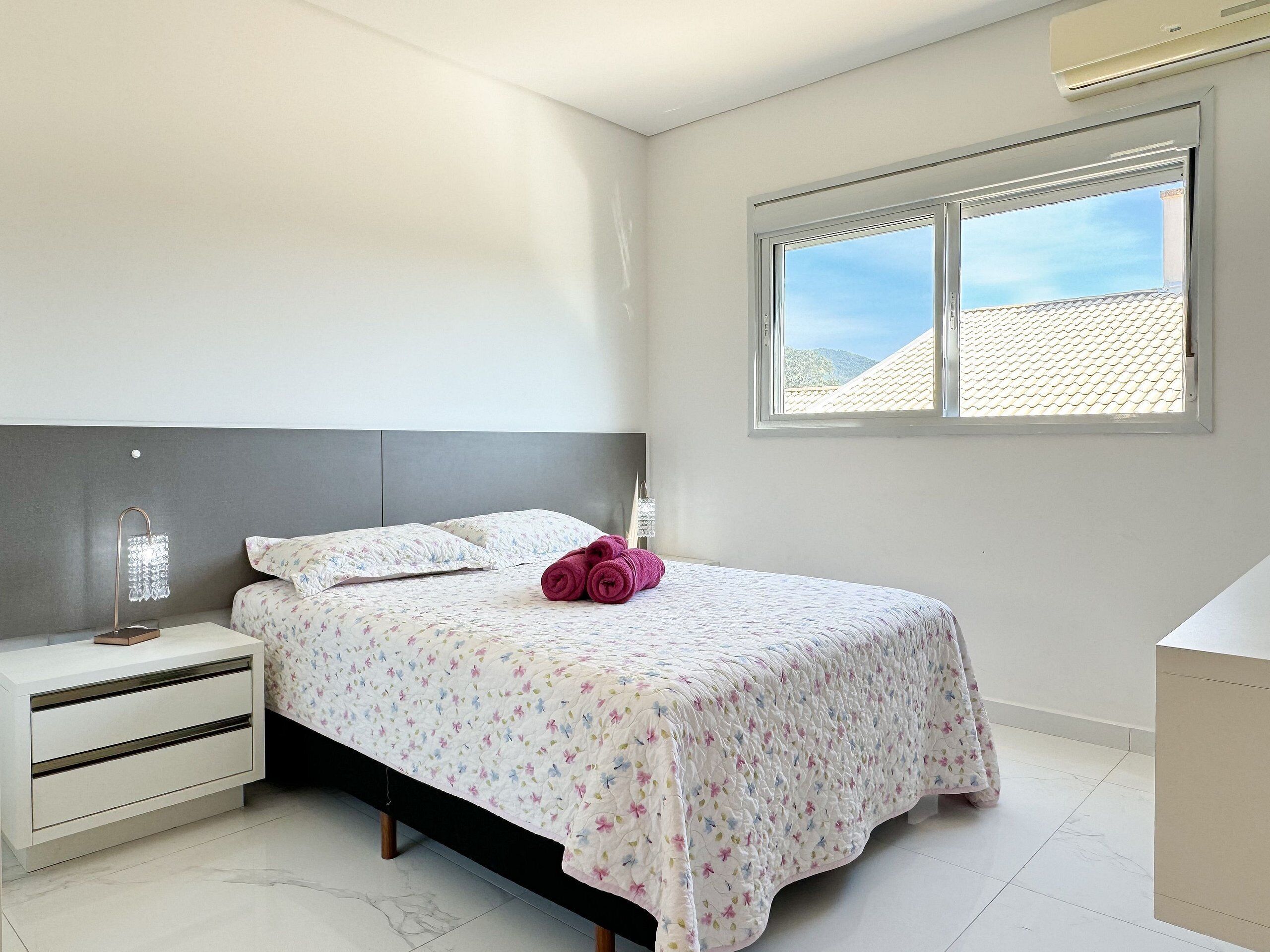 Un dormitorio con cama, mesita de noche, lámpara y ventana con vista al tejado.