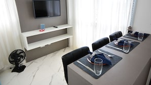 Une salle de conférence équipée d’un téléviseur à écran plat fixé au mur, d’une étagère blanche en dessous et d’une table à manger dressée avec des sets de table, des couverts et des verres.
