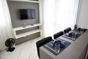 Un salón de conferencias con un televisor de pantalla plana montado en la pared, un estante blanco debajo y una mesa de comedor dispuesta con individuales, cubiertos y vasos.