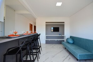 Una sala moderna con un sofá, un televisor montado en la pared y un comedor con sillas y una mesa dispuesta para dos personas.