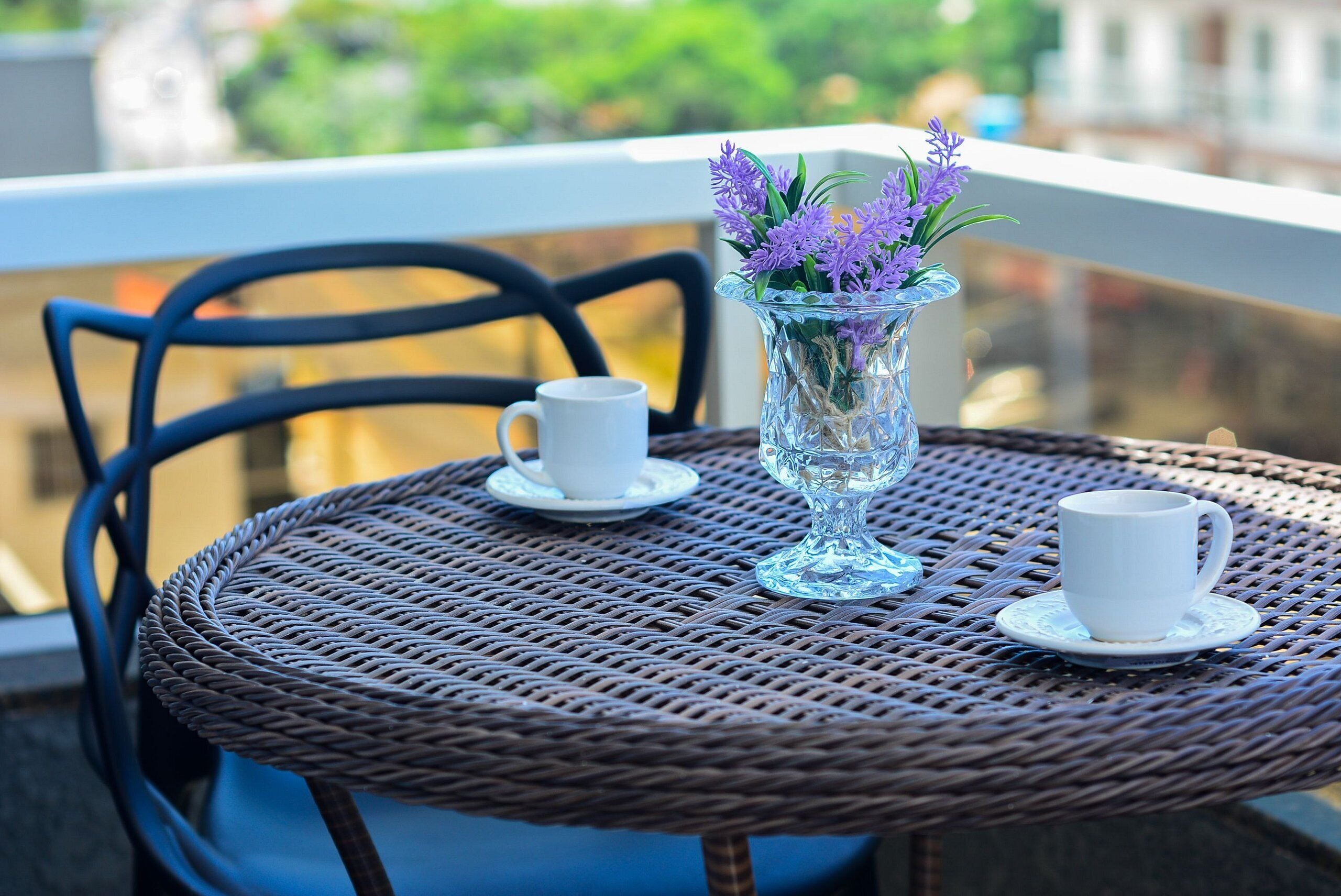 A round wicker table with two white cups and a vase with purple flowers on a balcony.