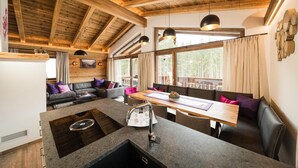 Un accogliente interno di una baita con soffitto in legno, una zona pranzo e un’area salotto con un divano e cuscini.