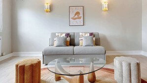 Una sala de estar con sofá, dos pufs, una mesa de centro de vidrio y una lámpara de pared.