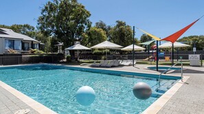 Piscina con sillas de descanso y sombrillas, rodeada de árboles y una zona vallada.
