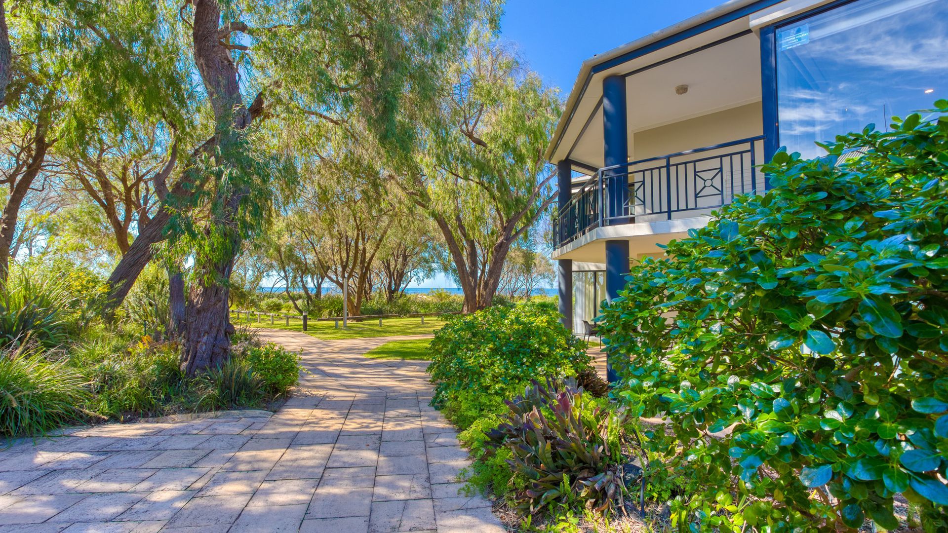 A pathway leading to a modern building with a balcony, surrounded by lush greenery and trees.