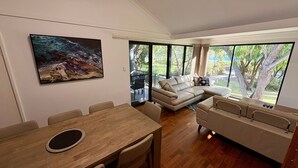 A modern living room with a wooden dining table, a beige sofa, and a large window offering a view of the outdoors.
