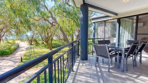 A balcony with a glass table and chairs, overlooking a garden with trees and a pathway.