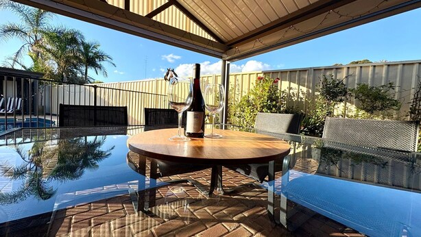 An outdoor dining area with a glass table, chairs, and a bottle of wine.