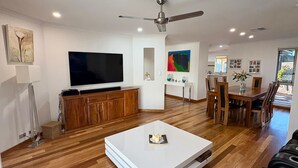 A modern living room with a flat-screen TV, wooden flooring, a ceiling fan, and a dining area with a table and chairs.