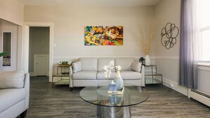 A living room with a glass coffee table, a sofa, a painting, and a vase with flowers.