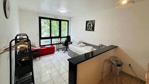 A modern hotel room with a large window, a desk, a chair, a bed, and a small kitchen area. - La Suite Buirette Hypercentre (Reims)