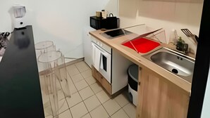 A compact kitchen with wooden cabinets, a sink, a microwave, and a small dining area with a chair. - La Suite Buirette Hypercentre (Reims)