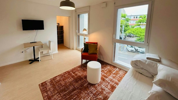 Une chambre d’hôtel moderne dotée d’une télévision à écran plat, d’un bureau, d’un fauteuil confortable, d’un canapé, d’une grande fenêtre offrant une vue sur la ville et d’un tapis à motifs.