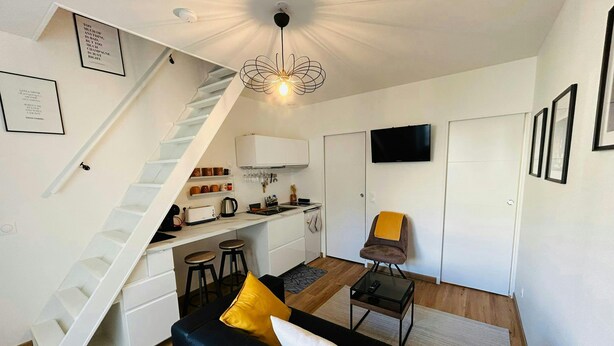 A compact living space with a kitchen, a dining area, a sofa with yellow cushions, a TV, and a staircase. - La Suite Marie Stuart Hypercentre (Reims)