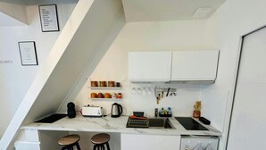 A compact kitchen with white cabinets, a built-in oven, a coffee maker, and a kettle. There are two stools and a small refrigerator. - La Suite Marie Stuart Hypercentre (Reims)