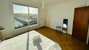 Une chambre avec une grande fenêtre, un lit recouvert d’une couverture blanche, un plancher en bois, un radiateur et une porte en bois.