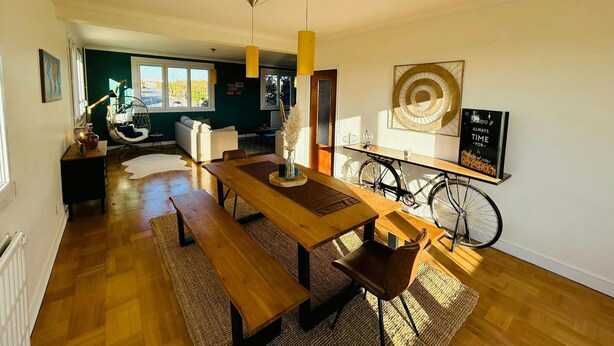 Un salon moderne avec une table à manger en bois, des bancs et un vélo. On aperçoit un canapé et un fauteuil suspendu à l’arrière-plan.