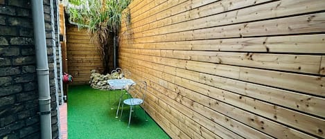 A narrow outdoor space with a green artificial turf, a metal table and chair, and a wooden fence.
