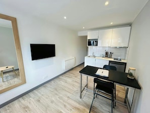 Eine moderne Küche mit weißen Schränken, einer Mikrowelle und einer Kaffeemaschine. Es gibt einen Esstisch mit Stühlen und einen großen Spiegel an der Wand.