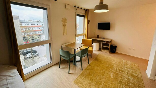 Un salon moderne doté d’une grande fenêtre, un coin repas avec une table et des chaises, un petit bureau avec un téléviseur et un luminaire suspendu.