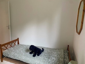 Ein kleines Zimmer mit einem Einzelbett, einem Holz-Kopfteil, einer gemusterten Bettdecke und einem dunkel gefärbten Stofftier auf dem Bett.