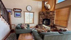 A cozy living room with a stone fireplace, a green sofa, a wooden coffee table, and a deer head mounted on the wall. - Rustic Mountain Lake Escape (Garden City)