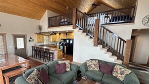 A spacious living area with a wooden ceiling, a staircase with a railing, and a kitchen in the background. - Rustic Mountain Lake Escape (Garden City)