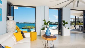 A modern living room with a white sofa, yellow cushions, and a large vase. There is a view of a pool and a beach through large windows. - Luxe Grand Villa Modern Spacious 6-bedroom with private Beach. Staff Included. (St. Ann)