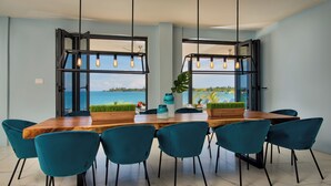 A dining area with a wooden table and teal chairs, large windows with black frames, and hanging light fixtures. - Luxe Grand Villa Modern Spacious 6-bedroom with private Beach. Staff Included. (St. Ann)