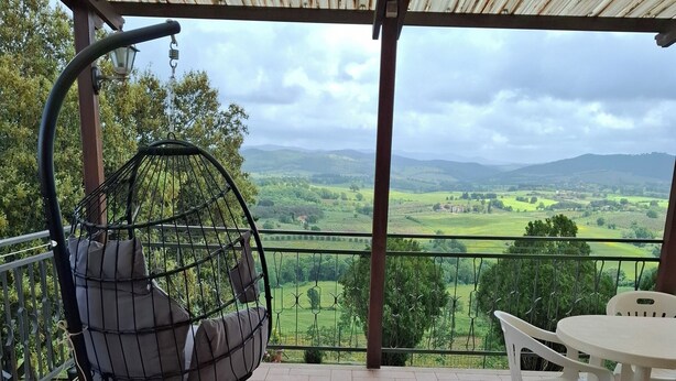 A covered patio with a hanging chair, a table with chairs, and a view of green fields and hills. - Vacation home Corrado 2 (Toscana)