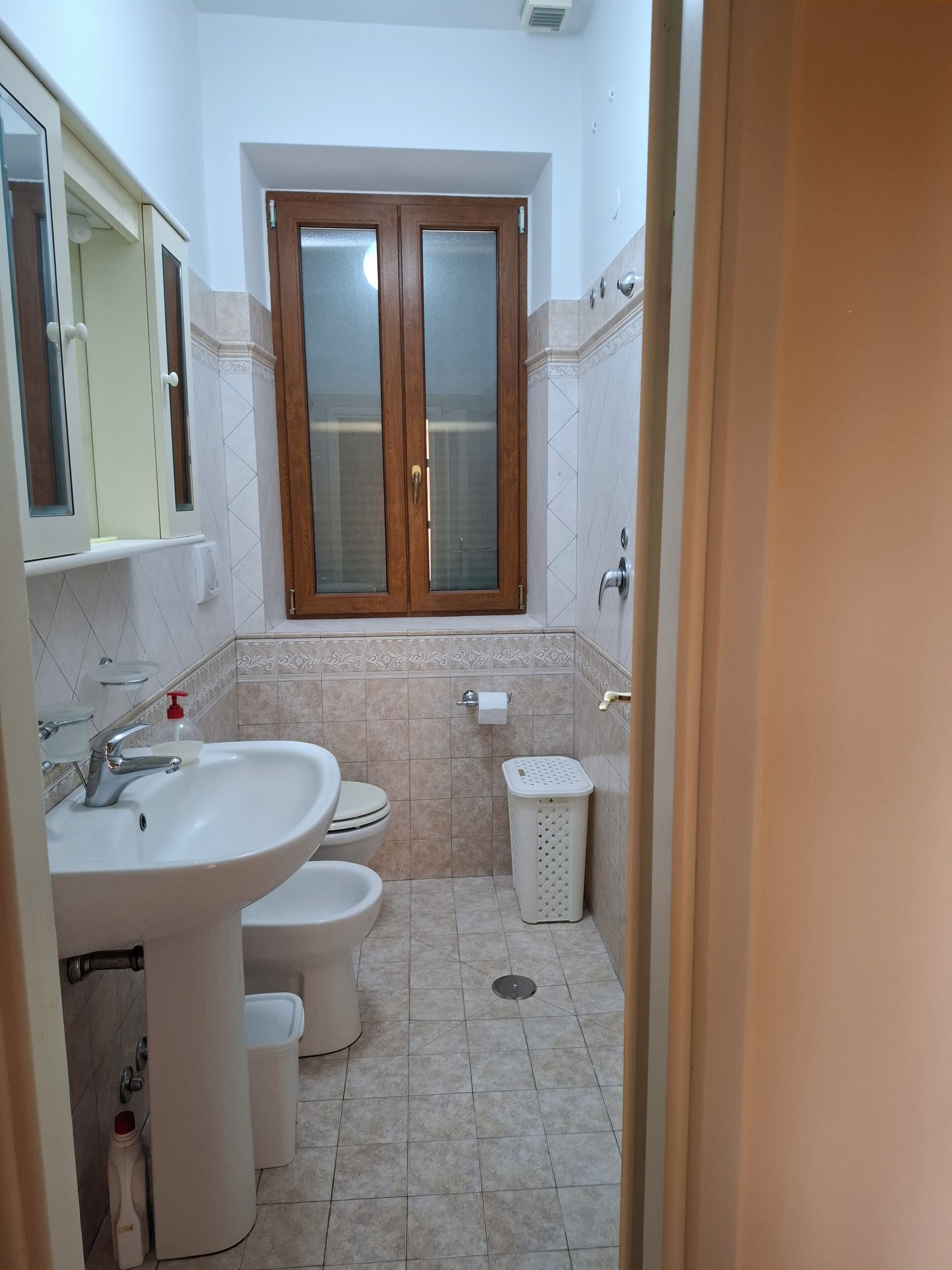 Une salle de bain avec un lavabo, deux toilettes, une fenêtre et une poubelle.